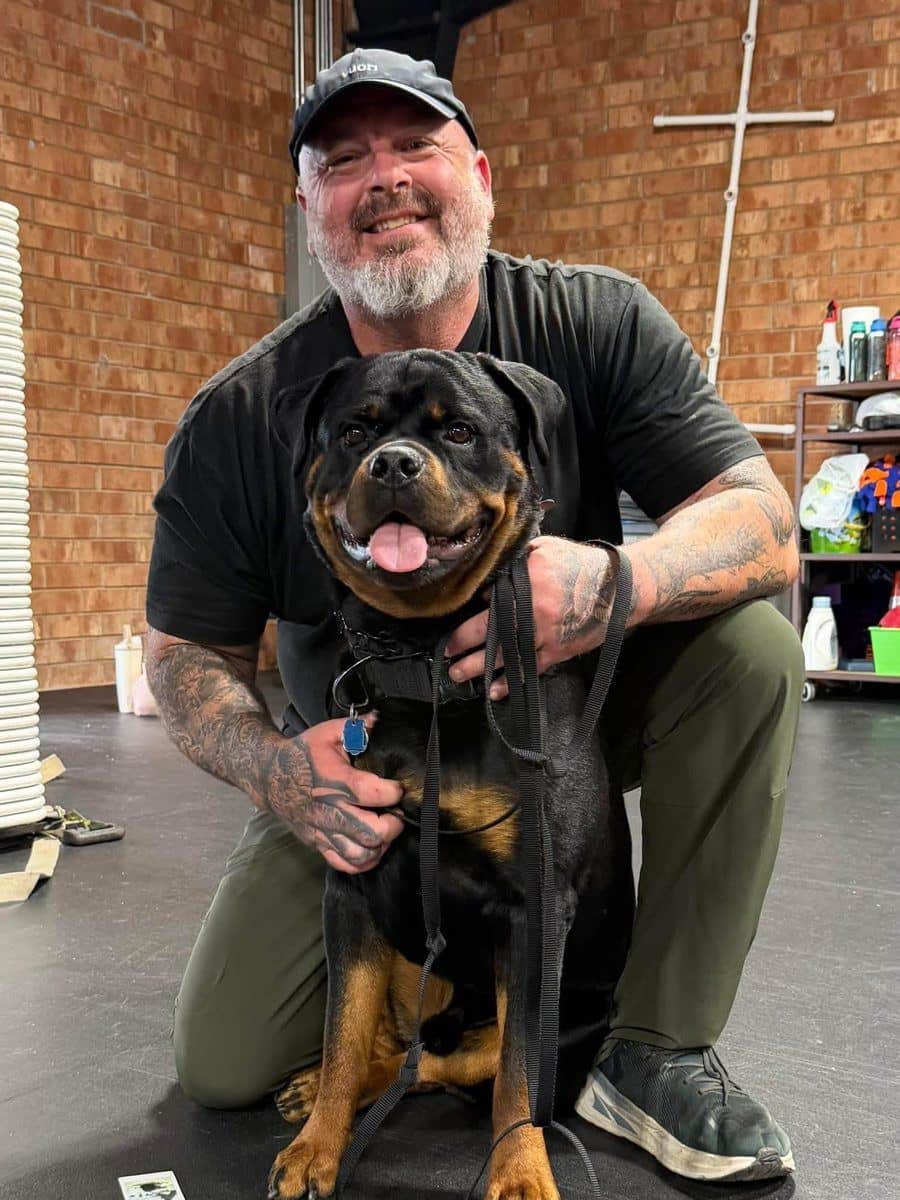 Larry Krohn kneeling beside a calm, happy Rottweiler on a leash in his training facility