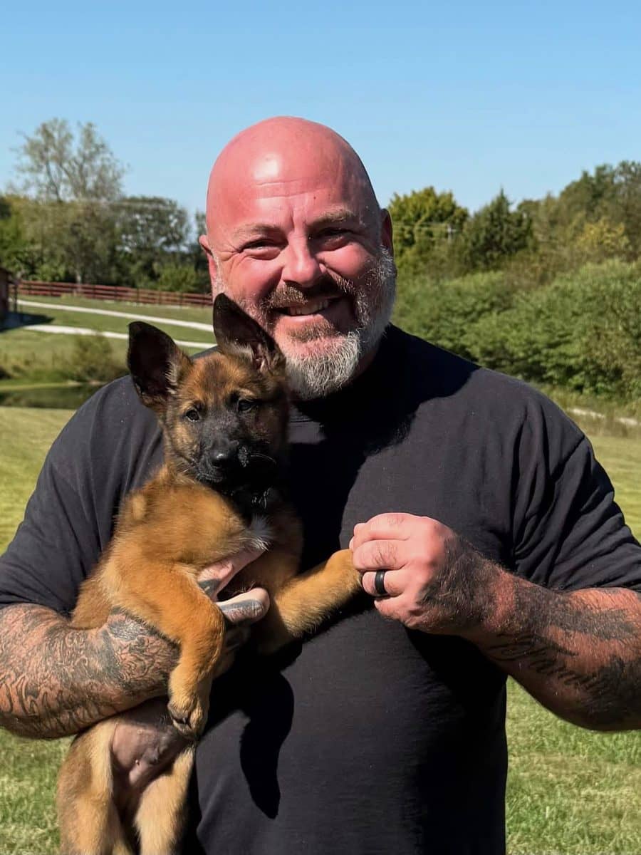 Larry Krohn smiling outdoors while holding a young Malinois puppy